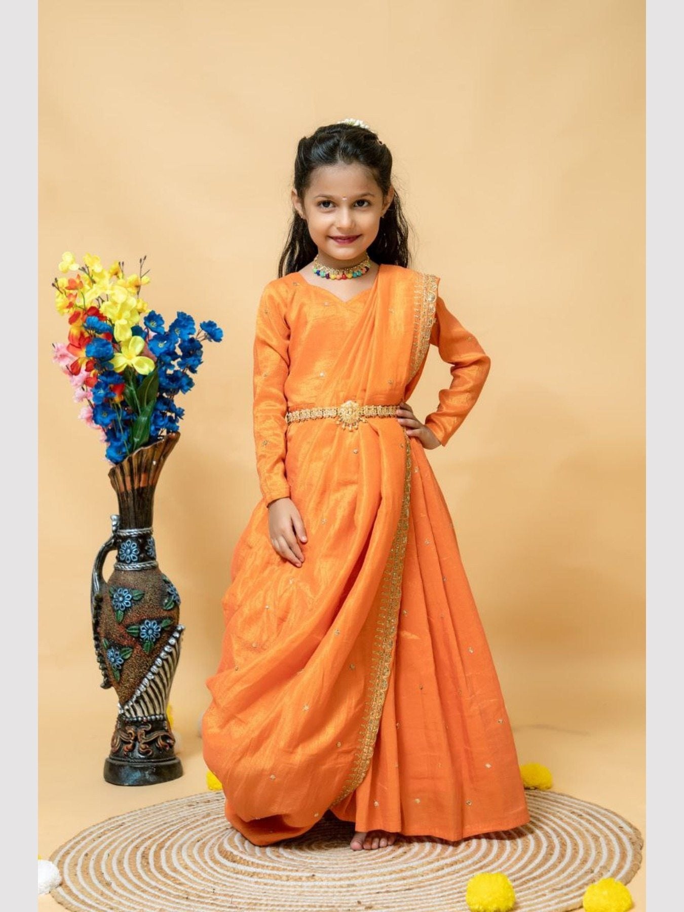 Girl in orange gown with dupatta, floral vase beside