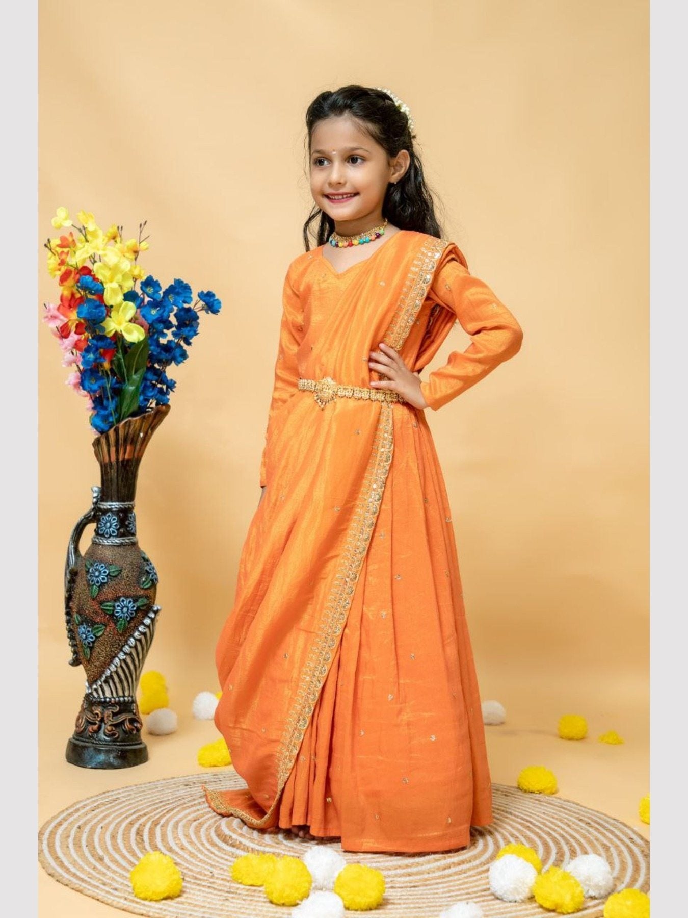 Girl smiling in orange silk gown with dupatta, floral vase